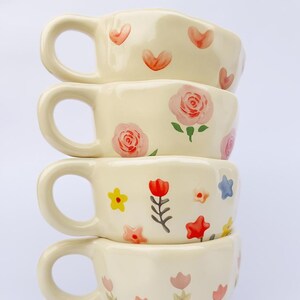 Può includere: Una pila di quattro tazze in ceramica color crema con disegni floreali dipinti a mano. Ogni tazza presenta un motivo diverso di fiori e cuori nei toni del rosso, rosa, giallo e blu. Le tazze hanno manici arrotondati.