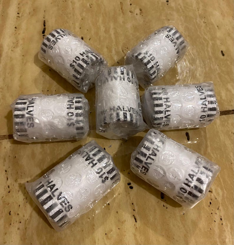 May include: Seven rolls of $10 Half Dollar coins, wrapped in paper and bubble wrap. The rolls are white with black text that reads "$10 HALVES". The coins are packaged for protection and display.