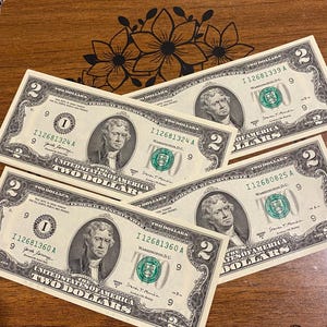 May include: Five United States two-dollar bills are displayed on a wooden surface. Each bill features a portrait of Thomas Jefferson and the words "Two Dollars." The bills are green and black with a white background.