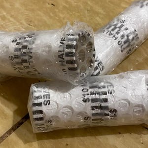 May include: Three rolls of $10 half dollars wrapped in bubble wrap. The rolls are white with black text that reads "$10 HALVES". The rolls are on a wooden surface.