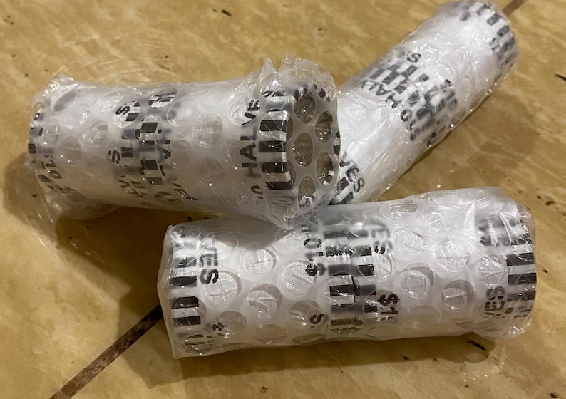 May include: Three rolls of $10 half dollars wrapped in bubble wrap. The rolls are white with black text that reads "$10 HALVES". The rolls are on a wooden surface.