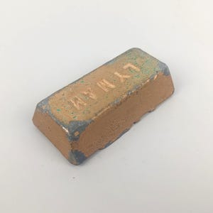 May include: A rectangular, brown polishing compound bar with the word "LYMAN" embossed on the top surface. The bar has a slightly weathered appearance with hints of green and grey around the edges.