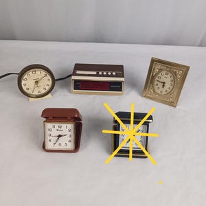 May include: A collection of vintage alarm clocks in various styles. Includes a round brass clock, a brown digital clock displaying "5:00", a gold framed clock, and two square clocks in brown and black.