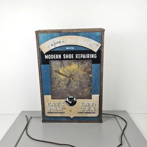Peut inclure: Ancienne enseigne publicitaire rectangulaire pour la réparation de chaussures. L'enseigne a un fond bleu avec le texte "Modern Shoe Repairing". L'enseigne comporte également le texte "Cat's Paw" et "Cat-Tex". L'enseigne a un cadre marron et un cadran d'horloge.