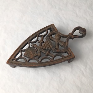 May include: A cast iron trivet with a decorative design. The trivet is triangular with a handle and features a shield, grapes, a figure, and other ornate details. The color is a rusty brown.