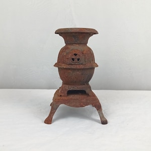 Puede incluir: Una pequeña estufa antigua de hierro fundido con un acabado marrón oxidado. La estufa tiene un cuerpo redondeado, una parte superior en forma de chimenea y tres patas curvas. Cuenta con una pequeña puerta con un diseño decorativo.