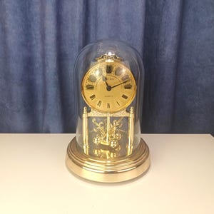 May include: A gold-tone Seiko quartz anniversary clock encased in a clear glass dome. The clock features a round gold face with black Roman numerals, black hands, and a rotating pendulum base with ornate gold-colored metal floral accents.
