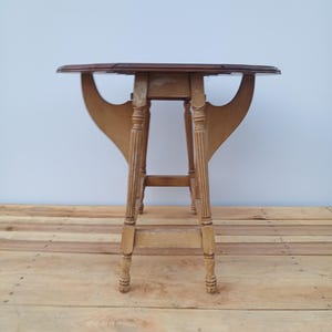 May include: A small, antique drop-leaf table with a dark brown tabletop and light brown, fluted legs. The table has a unique, curved support structure. The table is approximately 28 inches tall.