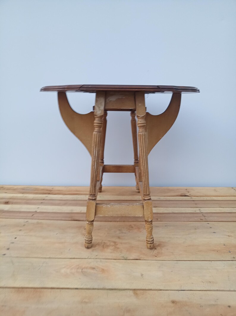 May include: A small, antique drop-leaf table with a dark brown tabletop and light brown, fluted legs. The table has a unique, curved support structure. The table is approximately 28 inches tall.