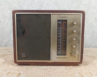 Może przedstawiać: Zabytkowe radio stereofoniczne AM/FM Realistic z ciemną drewnianą ramą. Radio posiada maskownicę głośnika, pokrętła strojenia i wyświetlacz częstotliwości. Płyta czołowa jest w kolorze jasnego złota z napisem "Realistic" na dole.