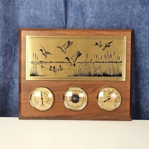 May include: A wooden weather station with a brass plate featuring a duck scene. The station includes a thermometer, barometer, and hygrometer. The thermometer displays temperatures in Fahrenheit.