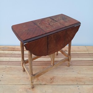 May include: A vintage drop-leaf table with a rich, reddish-brown finish. The table features a rectangular top with a patterned surface and rounded edges when the leaves are down. The legs are light-colored and have a fluted design. The table is approximately 30 inches long.