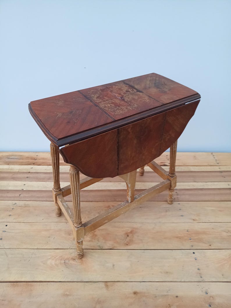 May include: A vintage drop-leaf table with a rich, reddish-brown finish. The table features a rectangular top with a patterned surface and rounded edges when the leaves are down. The legs are light-colored and have a fluted design. The table is approximately 30 inches long.