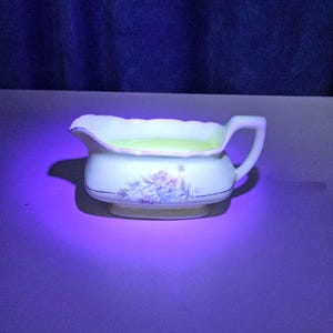 May include: A white ceramic gravy boat featuring a scalloped rim and a delicate floral pattern in shades of blue and pink. The interior of the vessel glows with a bright neon yellow hue under ultraviolet light, highlighting its unique composition.
