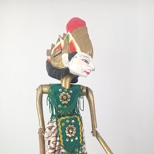 Puede incluir: Una marioneta tradicional indonesia Wayang Golek. La marioneta tiene una cara blanca con labios rojos, cabello negro y un tocado dorado con detalles rojos y amarillos. Viste una parte superior verde con adornos dorados y una falda estampada. Los brazos y las piernas son de madera.