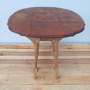 May include: A vintage, oval-shaped drop-leaf table with a dark wood finish. The table has a scalloped edge and rests on four ornate, light-colored legs. The table's dimensions are approximately 30 inches in diameter when open.