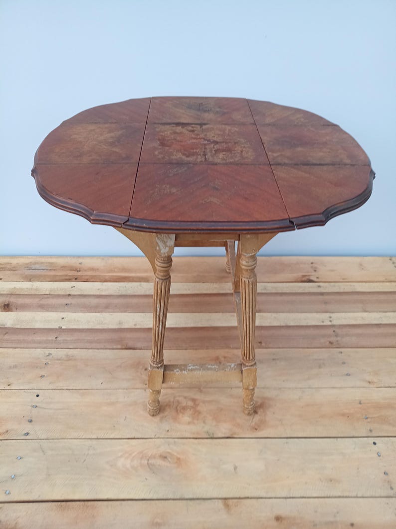 May include: A vintage, oval-shaped drop-leaf table with a dark wood finish. The table has a scalloped edge and rests on four ornate, light-colored legs. The table's dimensions are approximately 30 inches in diameter when open.