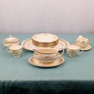 May include: A vintage cream-colored china set with gold trim and floral accents. The set includes cups, saucers, bowls, a serving platter, a gravy boat, and a sugar bowl with a lid. The dishes are arranged on a teal tablecloth.