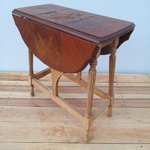 May include: A vintage wooden drop-leaf table with a rectangular top and rounded edges. The table has a dark brown tabletop and a light brown base with decorative legs. The table is partially folded down.