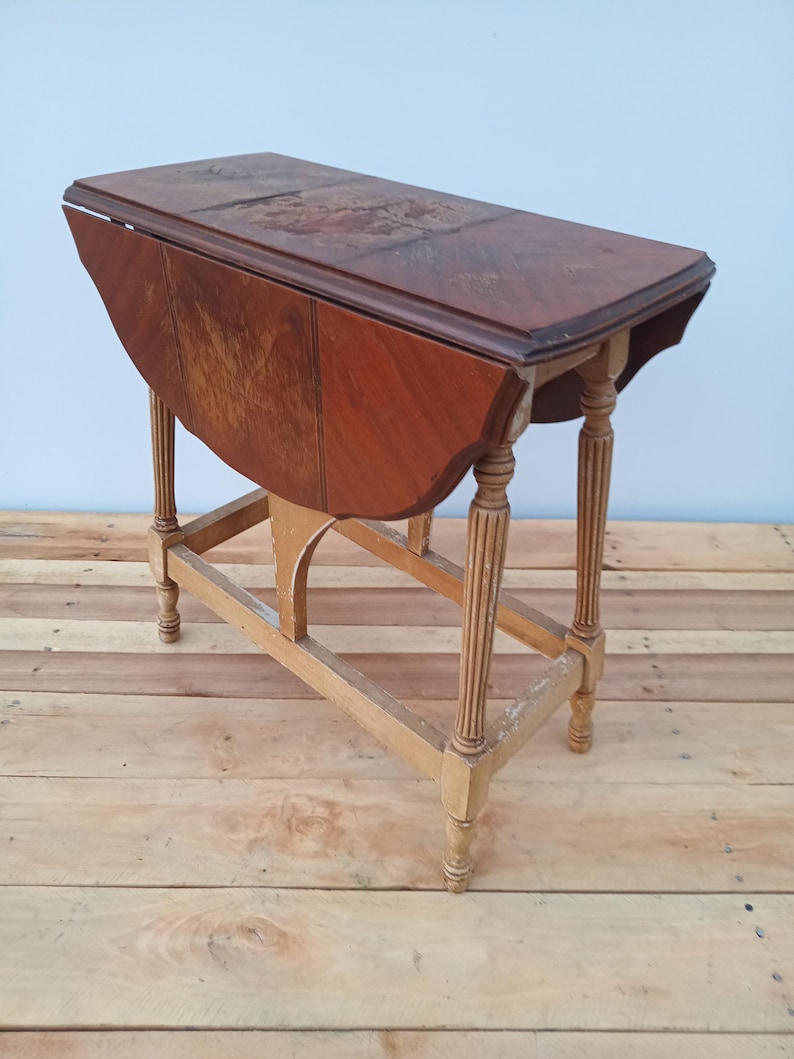 May include: A vintage wooden drop-leaf table with a rectangular top and rounded edges. The table has a dark brown tabletop and a light brown base with decorative legs. The table is partially folded down.