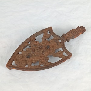 May include: A rusty brown cast iron trivet with a floral design. The trivet has a handle and a decorative openwork pattern. The handle has an orange accent. The trivet is designed to protect surfaces from hot items.