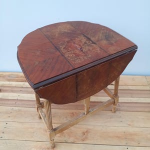 May include: A vintage, wooden drop-leaf table with a half-circle top. The tabletop is a rich, dark brown with a patterned design. The table has a light-colored, distressed wooden base with four legs. The table is approximately 30 inches long.