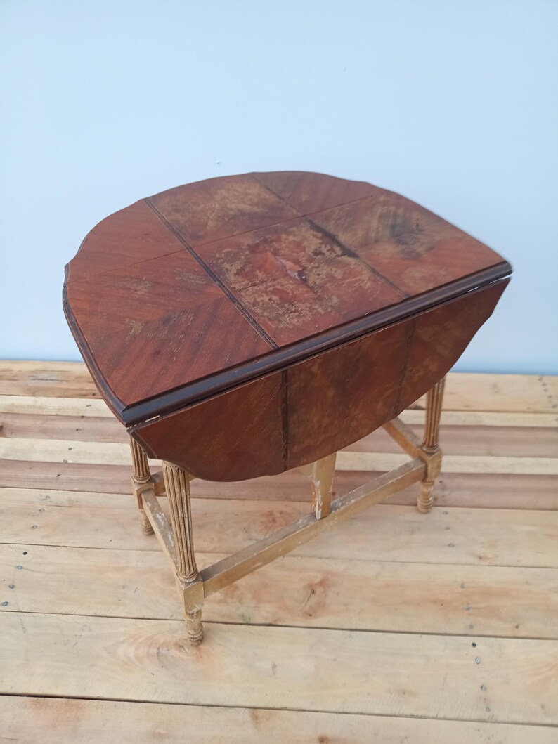 May include: A vintage, wooden drop-leaf table with a half-circle top. The tabletop is a rich, dark brown with a patterned design. The table has a light-colored, distressed wooden base with four legs. The table is approximately 30 inches long.