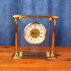 May include: A rectangular mantel clock featuring a gold-tone metal frame with two vertical pillars and a clear glass panel. The circular clock face has a white background, black Roman numerals, gold hands, and the word Quartz printed in black.