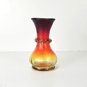 Puede incluir: Un florero de vidrio con un degradado de color que va desde el rojo intenso en el borde acampanado hasta el naranja brillante y amarillo en la base redondeada. El cuerpo tiene una textura en espiral y una banda de vidrio trenzado.
