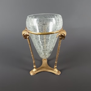 May include: A clear glass vase with a crackle pattern, supported by a gold-coloured metal stand. The stand features ram's head details and a circular band around the vase's midsection. The base has a three-footed design.