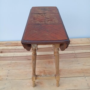 May include: A vintage, rectangular drop-leaf table with a dark wood grain top and decorative, fluted legs. The table has a warm, reddish-brown hue with some wear and tear visible on the surface. The legs are a lighter color with some paint chipping.