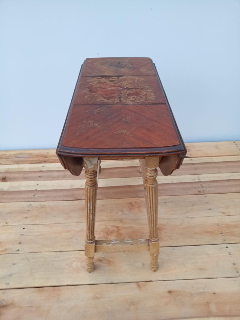 May include: A vintage, rectangular drop-leaf table with a dark wood grain top and decorative, fluted legs. The table has a warm, reddish-brown hue with some wear and tear visible on the surface. The legs are a lighter color with some paint chipping.