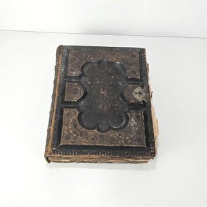 May include: An antique, rectangular book with a dark, weathered leather cover featuring intricate embossed patterns and a central scalloped panel. The spine has raised bands, and a small, tarnished metal clasp is attached to the right edge.