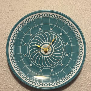 May include: A teal-colored decorative plate repurposed as a wall clock. The plate features a white geometric design with a central spiral and a border of circles. Gold-colored clock hands and a central mechanism complete the design.
