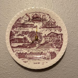 May include: A round, white decorative plate with a maroon illustration of buildings and text. The plate has a gold clock mechanism in the center and a braided edge. Text includes "International Bronze Bull Trophy".