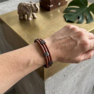 Könnte beinhalten: Ein mehrsträngiges Armband aus drei glatten, braunen Lederbändern, die durch gleichmäßig verteilte, rechteckige, dunkelmetallische Perlen zusammengehalten werden. Das Design besteht aus drei parallelen Lederbändern, die durch diese dunklen Metallakzente gesichert sind und ein strukturiertes, geschichtetes Erscheinungsbild am Handgelenk erzeugen.