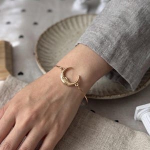 Könnte beinhalten: Ein goldfarbenes Metallarmband mit einem zentralen Halbmond-Anhänger, der ein detailliertes Gesicht aufweist. Das Armband besteht aus einem dünnen, glatten Metallreif, der an beiden Seiten mit dem Mondanhänger verbunden ist und einen Kreis bildet.