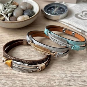 Könnte beinhalten: Drei Wickelarmbänder mit strukturierten Bändern in Braun, Hellgrau und Hellblau. Jedes Armband ist mit einem metallischen Federanhänger verziert, der durch farbige Fadenwicklungen befestigt ist. Das braune Armband hat eine silberne Feder, das graue eine goldene Feder und das hellblaue eine silberne Feder.