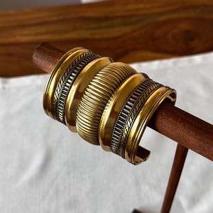 Könnte beinhalten: Eine Sammlung goldfarbener Metallarmbänder mit aufwendigen Designs, präsentiert auf einem Holzständer. Die Armbänder weisen verschiedene Texturen auf, darunter gerippte und gemusterte Details. Der Ständer ist aus dunklem Holz gefertigt, der Hintergrund ist neutral.