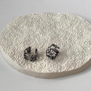 Könnte beinhalten: Zwei silberfarbene Ohrklemmen aus Metall mit einem komplizierten, durchbrochenen Filigran-Design und einem zentralen kreuzartigen Motiv. Die Schmuckstücke haben ein dunkles, oxidiertes Finish in den Vertiefungen, das die Rankenmuster betont.
