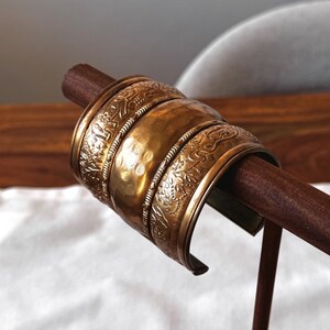 Könnte beinhalten: Ein Satz antiker goldfarbener Metallarmbänder mit aufwendigen geprägten Mustern. Die Armbänder werden auf einem Holzständer präsentiert, der ihre detaillierten Designs und ihr gealtertes Aussehen hervorhebt. Die Armbänder haben eine gehämmerten Textur.