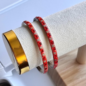 Könnte beinhalten: Zwei dünne rote Armreifen mit einem sich wiederholenden Muster aus kleinen quadratischen roten Emaille-Abschnitten, die sich mit strukturierten goldfarbenen Metallsegmenten abwechseln, neben einem glatten, polierten goldfarbenen Armreif auf einem weißen Samtständer.