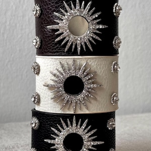 Könnte beinhalten: Ein Stapel aus drei breiten Armreifen in Dunkelbraun, Weiß und Schwarz mit strukturierter Oberfläche. Jeder Armreif hat ein zentrales, silberfarbenes Sonnenstrahlen-Emblem mit klaren Kristallen und kleinen Kristallnieten an den Seiten.