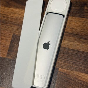 Op de afbeelding: Een zilveren afstandsbediening met een zwart Apple-logo, in een witte behuizing met een deksel. De behuizing heeft een cirkelvormige inkeping. De afstandsbediening ligt op een houten oppervlak.