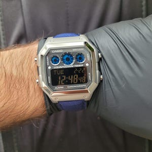 May include: A silver Casio digital watch with a blue fabric band. The watch face displays the time, date, and other functions. The watch has a square face with blue accents and the text "CASIO" and "WR100M".