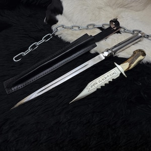 May include: A silver dagger with a serrated blade and a deer antler handle, resting on a black surface. The dagger is accompanied by a black leather sheath and a silver chain. The blade has etched text.