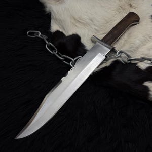 May include: A large, silver survival knife with a serrated edge and a dark wooden handle. A metal chain is draped over the knife, which rests on a black and white fur-like surface. The blade is sharp and angled.