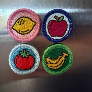 May include: Four embroidered patches featuring fruit illustrations. A yellow lemon on a pink background, a red apple on blue, a red tomato on light blue, and a yellow banana on green. Each patch is round with a stitched border.
