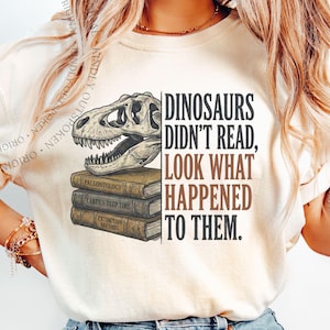 May include: An ivory t-shirt featuring a graphic of a dinosaur skull resting on a stack of three books labeled Paleontology, Earth's Deep Time, and Extinction Records, with the text Dinosaurs Didn't Read, Look What Happened To Them.