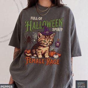 May include: A charcoal gray t-shirt featuring a graphic of a tabby kitten wearing a purple witch hat. The text reads FULL OF HALLOWEEN SPIRIT AND FEMALE RAGE. The design includes a spider, spiderwebs, pumpkins, and a small purple potion bottle.